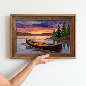 Pode incluir: Um painel retangular de vitral emoldurado em madeira, retratando uma canoa castanha a flutuar em águas calmas ao pôr do sol. A cena apresenta um céu laranja e roxo vibrante, pinheiros verde-escuros na margem e reflexos na água.