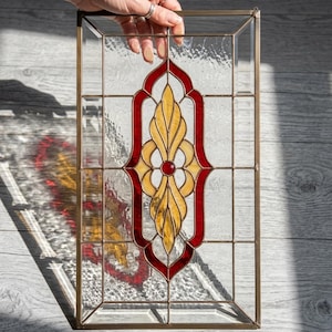 Puede incluir: Panel rectangular de vidriera con marco de latón. El diseño presenta un motivo central de elementos florales estilizados amarillos y rojos, con un pequeño círculo rojo en el centro. El vidrio tiene una apariencia texturizada.