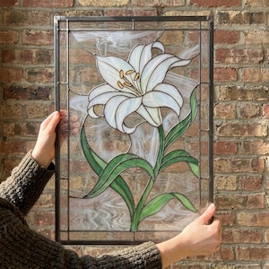 May include: A rectangular stained glass panel featuring a large white lily flower with yellow stamens and green leaves. The glass has a swirled, translucent texture set within a dark metal frame, showcasing a floral design.