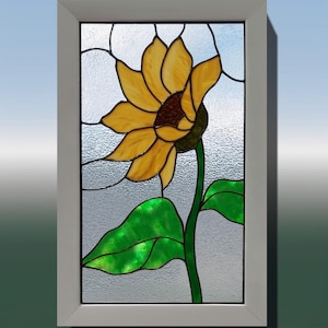 May include: Stained glass window panel featuring a vibrant sunflower. The sunflower has yellow petals, a brown center, and a green stem and leaves. The panel is framed in a white border, creating a decorative piece for home decor.