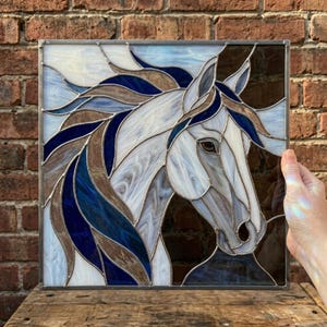 Op de afbeelding: Gebrandschilderd glaspaneel met een paardenkop. Het kunstwerk gebruikt tinten wit, blauw en bruin om de manen en het gezicht van het paard weer te geven. Het paneel heeft een metalen rand.