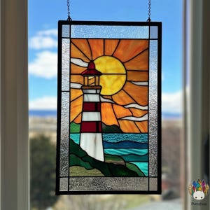 May include: Stained glass panel depicting a lighthouse with red and white stripes, a red roof, and a bright yellow sun. The scene includes blue water, green land, and a blue sky. The panel is framed in black and hangs by a chain.