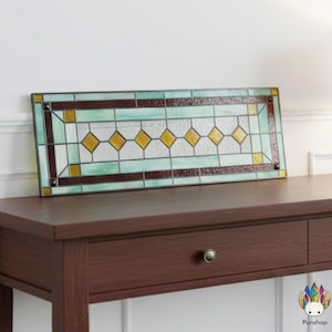 May include: Rectangular stained glass panel with a geometric design. The panel features a border of brown and green, with yellow diamond shapes in the center. The glass is set in a black frame. The panel is resting on a dark brown wooden table.