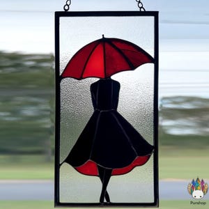 Puede incluir: Panel de vidriera con la silueta de una persona bajo un paraguas rojo. La persona lleva un vestido negro con un dobladillo rojo. El panel es rectangular con un borde negro y un fondo de vidrio texturizado.