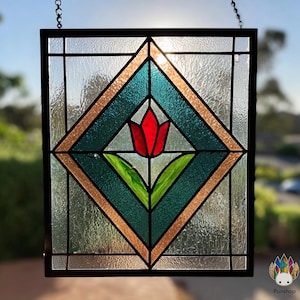 May include: Stained glass panel featuring a red tulip flower with green leaves, set within a teal and gold diamond design. The panel is framed in black and hangs from a chain. The background is blurred, suggesting an outdoor setting.