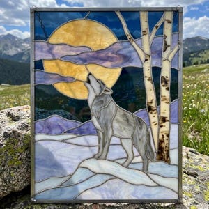 Op de afbeelding: Een rechthoekig glas-in-loodpaneel met een grijze wolf die naar een grote gele maan huilt. De scène bevat witte sneeuw, paarse wolken en twee berkenbomen met donkere markeringen, alles gevat in een metalen frame met zichtbare soldeernaden.