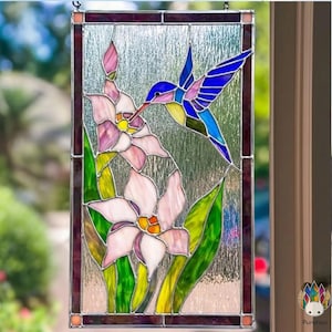 Pode incluir: Painel de vitral com um beija-flor e flores. O beija-flor é retratado com tons de azul, rosa e amarelo, pairando perto de flores rosa e brancas com folhas verdes. O painel tem uma borda marrom escura.