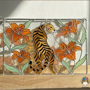 Pode incluir: Painel de vitral com um tigre com listras laranja e pretas, rodeado por lírios laranja e folhas verdes. A obra de arte é emoldurada com uma borda de metal prateado, criando uma peça decorativa.