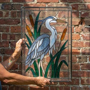 Puede incluir: Un panel rectangular de vidriera que muestra una garza azul y blanca entre juncos verdes y espadañas de color ámbar. La obra está construida con líneas de plomo metálico oscuro que forman un patrón de cuadrícula alrededor del ave.