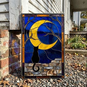 May include: A rectangular stained glass panel featuring a black cat silhouette sitting on a stone wall, looking up at a large yellow crescent moon. The background is a deep blue night sky with bare tree branches, framed by a dark border.