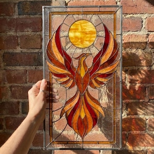 May include: A rectangular stained glass panel featuring a central phoenix with outstretched wings in shades of red, orange, and yellow. A circular yellow sun sits above the phoenix, all encased in a dark metal frame with clear glass borders.