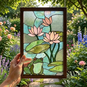 May include: A rectangular stained glass panel in a dark wood frame featuring a green frog sitting among light green lily pads and three light pink lotus flowers against a textured light blue background with visible lead lines.