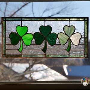 May include: Stained glass window featuring three shamrocks in shades of green. The shamrocks are set against a textured, clear background, framed by a dark border. A decorative piece for St. Patrick's Day.