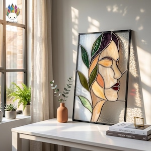Pode incluir: Painel de vitral com o perfil de uma mulher e um design de folhas. A obra utiliza tons de verde, laranja, amarelo e roxo. Emoldurado em preto, repousa sobre uma mesa branca. Um pequeno vaso de terracota e livros também estão presentes.