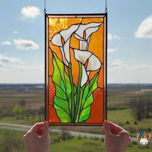 May include: Stained glass panel featuring white calla lilies with green leaves against an orange background. The panel is framed in black and hangs from a chain. The artwork is held up against a backdrop of a blue sky and a field.