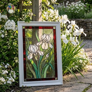May include: A framed stained glass panel featuring white iris flowers with green leaves and stems. The panel has a white frame with a red border. The artwork is displayed outdoors, surrounded by greenery and white flowers.