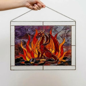 May include: A rectangular stained glass panel featuring a central red dragon surrounded by vibrant orange and yellow flames. The dragon rests on dark, textured glass rocks against a purple and grey background, all framed by clear glass and a metal chain.