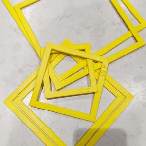 May include: A set of four yellow plastic frames, each with a different size and shape. The frames are stacked on top of each other and have the words "N.I.E." and "A.I.N." printed on them.