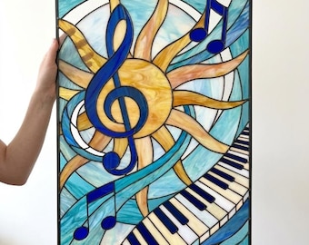 May include: A rectangular stained glass panel featuring a central yellow sun with radiating rays, a prominent blue treble clef, floating blue musical notes, and a diagonal piano keyboard at the bottom, all set against a light blue background.