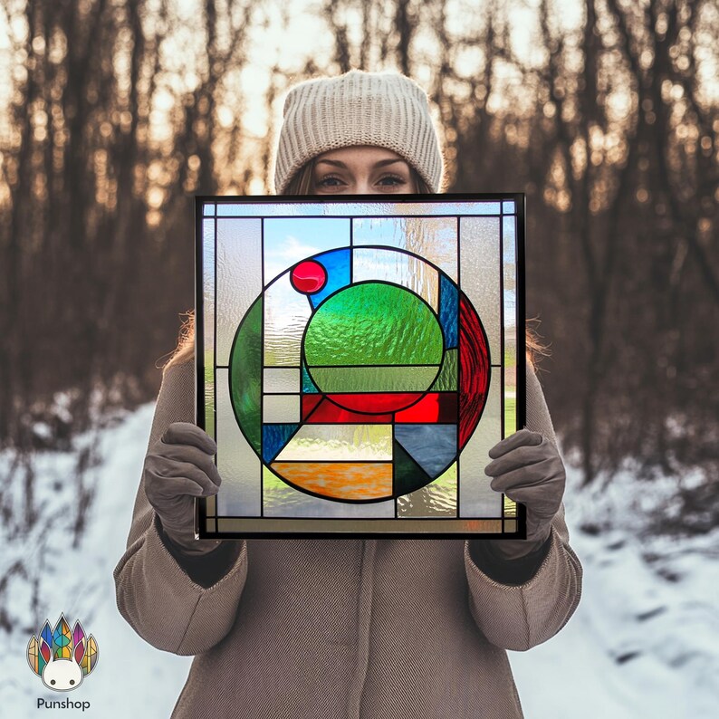Pu&ograve; includere: Un pannello di vetro colorato con un design geometrico che presenta un cerchio con un centro verde, accenti rossi, blu e gialli. Il pannello &egrave; incorniciato in nero.