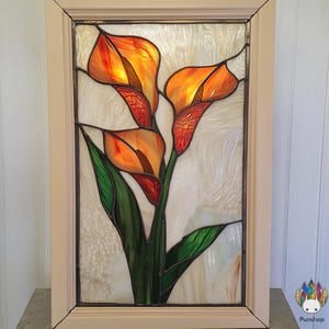 Op de afbeelding: Een glas-in-loodraam met drie oranje calla lelies met groene bladeren. Het paneel is ingelijst in een licht houten frame.