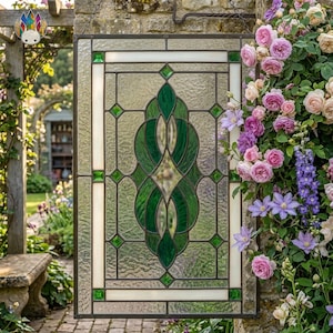 May include: A rectangular stained glass window with a green and white design. The central motif is a stylized green leaf pattern, framed by white and green geometric shapes. The window is hanging outdoors, with a garden and flowers in the background.