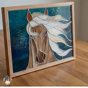 May include: Stained glass art piece featuring a horse's head with flowing mane. The artwork uses shades of brown, white, and tan against a teal background. The piece is framed in a light-colored wood frame, showcasing the intricate details of the design.