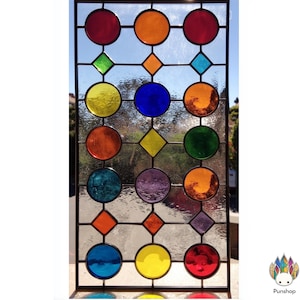 Könnte beinhalten: Ein rechteckiges Buntglasfenster mit einem schwarzen Metallrahmen. Das Design zeigt ein Gitter aus farbigen Kreisen und Rauten in Rot-, Orange-, Gelb-, Grün-, Blau- und Lilatönen. Das Fenster ist transparent und lässt Licht durch.