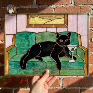 May include: Stained glass artwork depicting a black cat with a martini on a green sofa. The piece has a gold frame, with pink and yellow sections. The brand name "Punshop" is visible. The artwork is suitable for home decor.