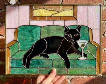 Puede incluir: Vidriera que representa un gato negro en un sofá verde, sosteniendo un martini. La obra de arte tiene un marco dorado y detalles en rosa y amarillo. La marca "Punshop" es visible. Ideal para la decoración del hogar.
