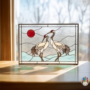 Peut inclure: Panneau en vitrail représentant deux grues se faisant face, avec un soleil rouge en arrière-plan. Les oiseaux ont des plumes blanches et marron, avec des accents rouges sur la tête. Le panneau est encadré de métal foncé.