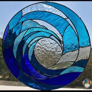 May include: Circular stained glass window depicting a stylized ocean wave. The design features shades of blue and clear glass, creating a swirling pattern. The glass has textured and smooth sections, with black lines separating the pieces.