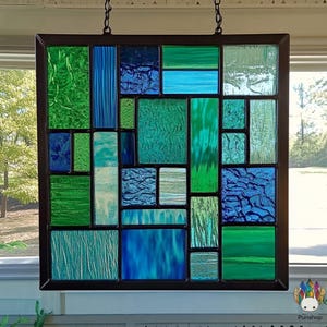 May include: A stained glass window panel with a geometric design featuring shades of blue and green. The panel is framed in black and is hung on a wall.