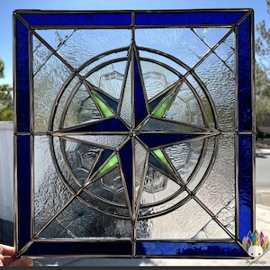 May include: A square stained glass window with a compass rose design. The window has a dark blue border and a clear glass background. The compass rose is blue and green. The intricate design is suitable for home decor, with measurements in centimetres.