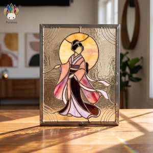 Op de afbeelding: Gebrandschilderd glaspaneel met een geisha in kimono, tegen een gouden achtergrond. Het kunstwerk gebruikt roze, paarse en bruine tinten, omlijst door een metalen rand. Een decoratief stuk.