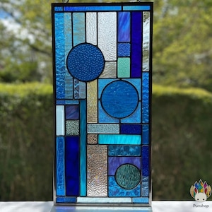 Op de afbeelding: Een rechthoekig glas-in-loodpaneel met een geometrisch ontwerp. Het paneel heeft verschillende tinten blauw, waaronder hemelsblauw, azuur en marineblauw, gerangschikt in rechthoekige en cirkelvormige vormen. Het glas heeft getextureerde en gladde secties.