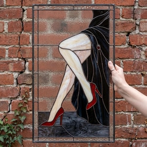 Puede incluir: Panel de vidrieras que representa las piernas de una mujer con un vestido negro y tacones rojos. La obra de arte presenta un fondo oscuro y un marco de metal decorativo. El panel se sostiene contra una pared de ladrillos.