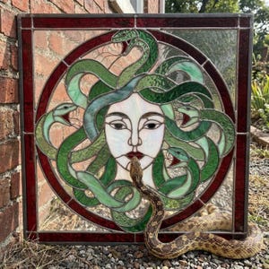 May include: A square stained glass panel featuring a central face surrounded by green glass snakes within a circular frame, bordered by deep red glass. A live, tan-patterned snake rests in front of the panel, mirroring the imagery of the glass art.