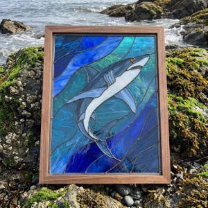 May include: A rectangular stained glass panel featuring a shark swimming through deep blue and turquoise water, encased in a simple wooden frame. The shark is rendered in shades of grey and white with a single dark eye, set against wavy glass.