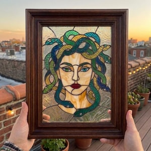 May include: A stained glass panel in a dark wood frame depicting a face with green and yellow snakes for hair. The figure has light skin, dark red lips, and heterochromatic eyes, one green and one blue, set against a textured clear glass background.