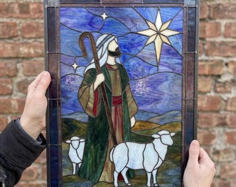 May include: A rectangular stained glass panel featuring a shepherd in a green robe holding a wooden staff, accompanied by two white sheep. The background depicts a dark blue night sky with a large, bright, multi-pointed star above.