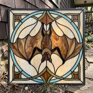 Op de afbeelding: Een vierkant glas-in-loodpaneel met een centrale vleermuis met uitgestrekte vleugels in bruintinten, gevat in een cirkelvormig frame van lichtblauw en crèmekleurig glas. Het ontwerp wordt bijeengehouden door donkere metalen loodlijnen.