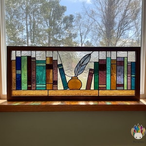 Può includere: Pannello in vetro colorato raffigurante una libreria con libri colorati, una penna e un calamaio. Il pannello è incorniciato in legno scuro e posto davanti a una finestra con vista sugli alberi. L'opera d'arte presenta vari colori, tra cui blu, verde e rosso.