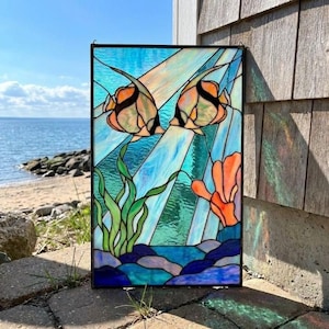 Peut inclure: Un panneau rectangulaire en vitrail représentant deux poissons orange et noirs nageant parmi des algues vertes et du corail orange. Le fond est composé de segments de verre bleu clair et blanc créant un effet sous-marin avec des rochers bleus.