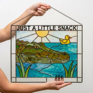 Könnte beinhalten: Ein rechteckiger Buntglas-Sonnenfänger mit einem grünen Alligator im blauen Wasser, einer gelben Ente und einer Sonne. Der Text JUST A LITTLE SNACK steht oben. Das Stück ist metallgerahmt und verfügt über eine Kette zum Aufhängen des Objekts.