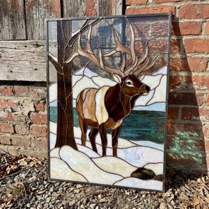 May include: A rectangular stained glass panel featuring an elk standing in a snowy landscape. The elk has brown and tan glass segments with large antlers. A dark brown tree trunk stands to the left, with a deep green river and white snow.