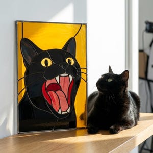Könnte beinhalten: Ein Buntglasfenster mit einer schwarzen Katze mit offenem Maul und gelben Augen vor gelbem Hintergrund. Eine echte schwarze Katze sitzt neben dem Panel auf einer Holzoberfläche.