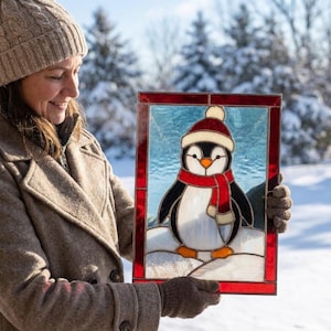 May include: A rectangular stained glass panel featuring a penguin wearing a red and white hat and scarf. The penguin stands on a white snowy base against a light blue background, all framed by a deep red border with dark metal solder lines.
