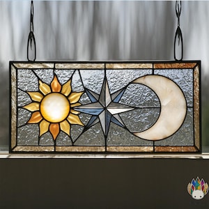 Può includere: Pannello in vetro colorato con elementi celesti. Il design include un sole giallo e arancione, una stella blu e grigia e una luna crescente bianca. Il pannello è rettangolare con una cornice in metallo nero e catene di sospensione.