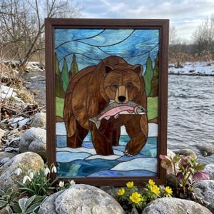Peut inclure: Un panneau rectangulaire en vitrail dans un cadre en bois foncé représentant un ours brun debout dans une rivière bleue et blanche, tenant un poisson rose et argenté dans sa gueule. Le fond montre des arbres verts sous un ciel bleu tourbillonnant.